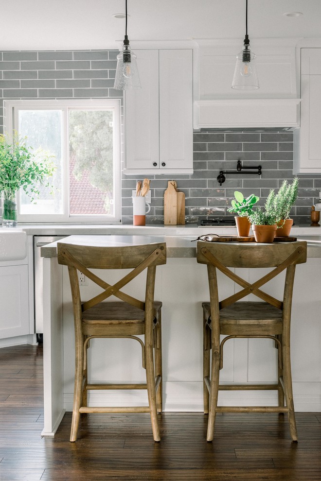 Example of a mid-sized farmhouse single-wall laminate floor and brown floor open concept kitchen design in San Diego with a farmhouse sink, shaker cabinets, white cabinets, quartz countertops, gray backsplash, ceramic backsplash, stainless steel appliances, an island and gray countertops