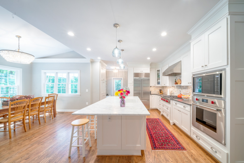Photo of a medium sized classic kitchen/diner in DC Metro with a built-in sink, white cabinets, granite worktops, beige splashback, stainless steel appliances, light hardwood flooring, an island, beige floors, white worktops and a vaulted ceiling.