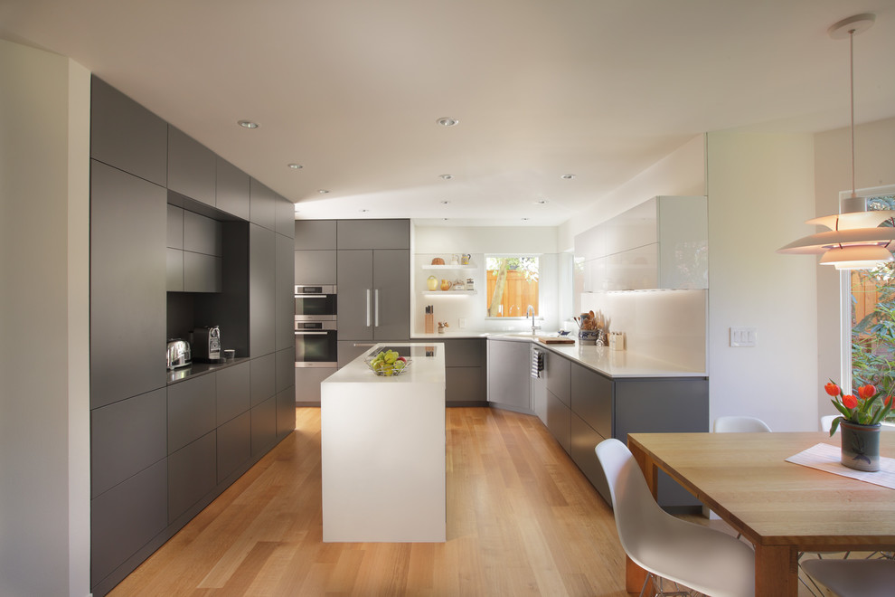 Moderne Wohnküche mit flächenbündigen Schrankfronten, grauen Schränken, Küchenrückwand in Weiß, Küchengeräten aus Edelstahl, hellem Holzboden und Kücheninsel in Portland