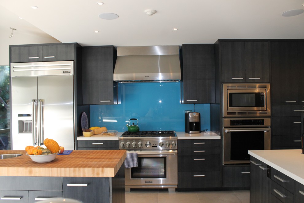 サンフランシスコにあるモダンスタイルのおしゃれなキッチン (ダブルシンク、フラットパネル扉のキャビネット、中間色木目調キャビネット、クオーツストーンカウンター、青いキッチンパネル、ガラス板のキッチンパネル、シルバーの調理設備、磁器タイルの床) の写真