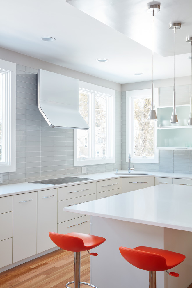 Design ideas for a medium sized contemporary u-shaped open plan kitchen in Boston with a single-bowl sink, flat-panel cabinets, white cabinets, engineered stone countertops, glass tiled splashback, white appliances, medium hardwood flooring, an island, grey splashback and white worktops.