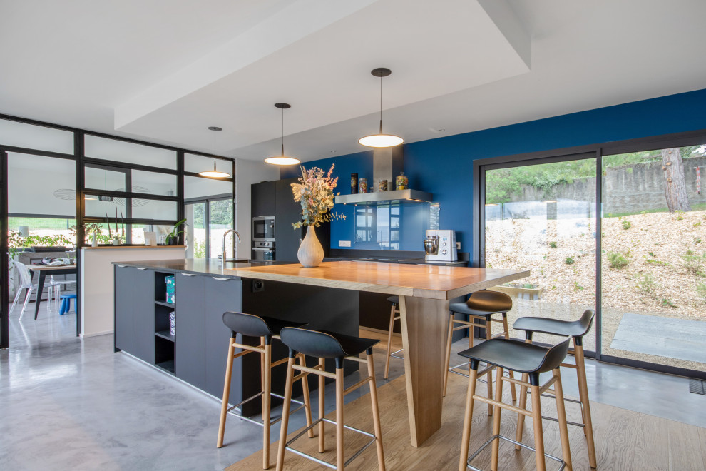 На фото: кухня в современном стиле с плоскими фасадами, черными фасадами, синим фартуком, фартуком из стекла, бетонным полом, островом, серым полом и черной столешницей