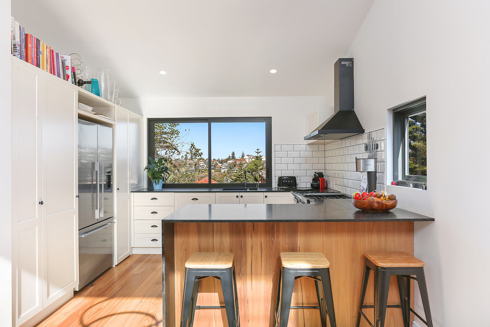 Contemporary u-shaped kitchen in Sydney with a double-bowl sink, shaker cabinets, white cabinets, white splashback, metro tiled splashback, stainless steel appliances, light hardwood flooring, a breakfast bar and black worktops.