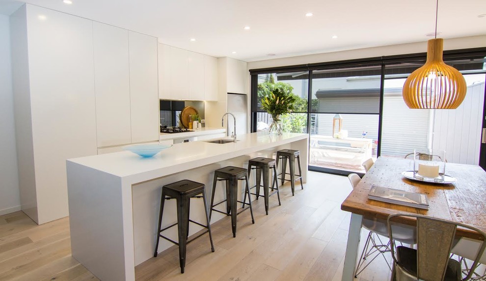 This is an example of a medium sized contemporary single-wall kitchen/diner in Melbourne with a single-bowl sink, flat-panel cabinets, white cabinets, engineered stone countertops, black splashback, glass sheet splashback, white appliances, light hardwood flooring and an island.