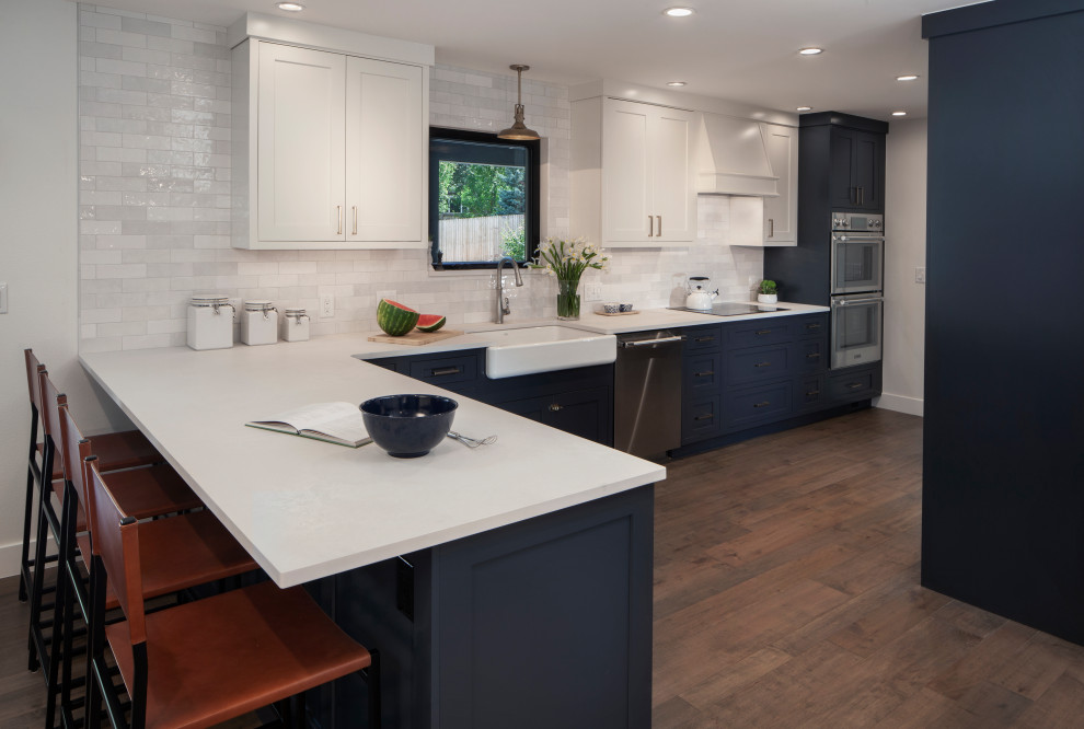 Photo of a country l-shaped kitchen/diner in Denver with a belfast sink, shaker cabinets, blue cabinets, engineered stone countertops, white splashback, ceramic splashback, stainless steel appliances, medium hardwood flooring, a breakfast bar and white worktops.