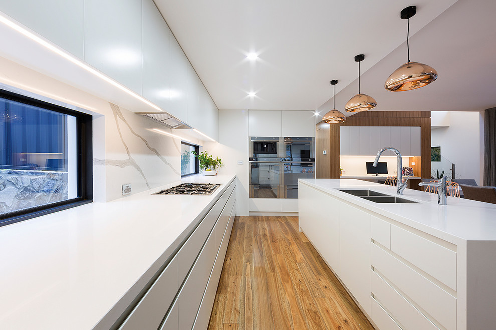 Modern inredning av ett stort kök, med släta luckor, vita skåp, vitt stänkskydd, rostfria vitvaror, en köksö, brunt golv, en undermonterad diskho, fönster som stänkskydd och mellanmörkt trägolv