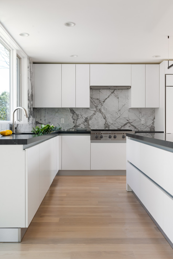 Photo of a contemporary l-shaped kitchen in Boston with a submerged sink, flat-panel cabinets, white cabinets, grey splashback, stone slab splashback, stainless steel appliances, light hardwood flooring, an island, beige floors and black worktops.