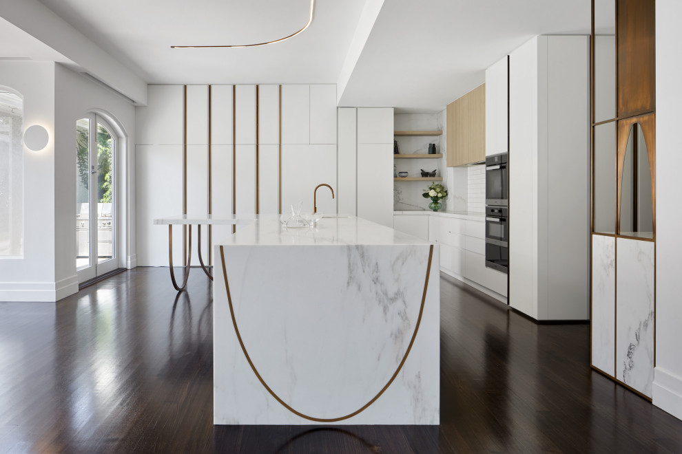 Design ideas for a large contemporary l-shaped kitchen/diner in Melbourne with a submerged sink, white cabinets, marble worktops, white splashback, ceramic splashback, black appliances, medium hardwood flooring, an island, brown floors and white worktops.