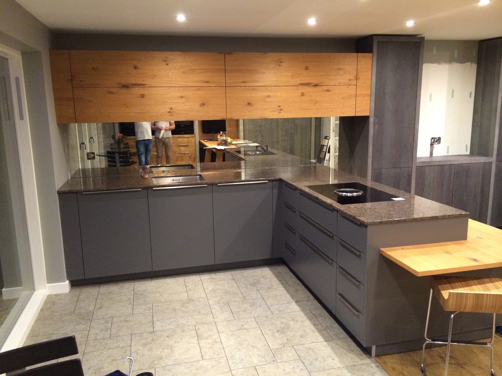 Example of a mid-sized urban l-shaped ceramic tile eat-in kitchen design in Cambridgeshire with a drop-in sink, flat-panel cabinets, gray cabinets, granite countertops, metallic backsplash, glass sheet backsplash, black appliances and no island