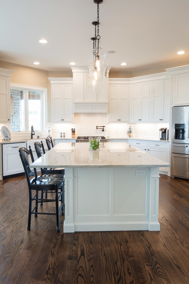 Photo of a large classic u-shaped kitchen/diner in Cincinnati with a belfast sink, shaker cabinets, white cabinets, marble worktops, white splashback, glass tiled splashback, stainless steel appliances, medium hardwood flooring, an island and brown floors.