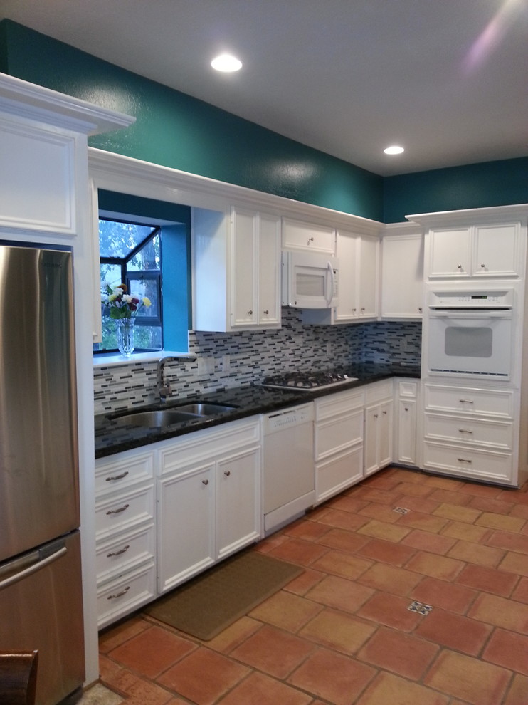 Mid-sized transitional terra-cotta tile kitchen photo in Houston with a double-bowl sink, white cabinets, granite countertops, blue backsplash, glass tile backsplash, white appliances and a peninsula