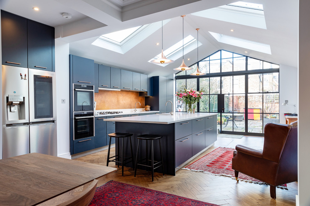 Große, Zweizeilige Moderne Wohnküche mit flächenbündigen Schrankfronten, Kücheninsel, Unterbauwaschbecken, blauen Schränken, Küchenrückwand in Orange, Küchengeräten aus Edelstahl, braunem Boden und weißer Arbeitsplatte in London