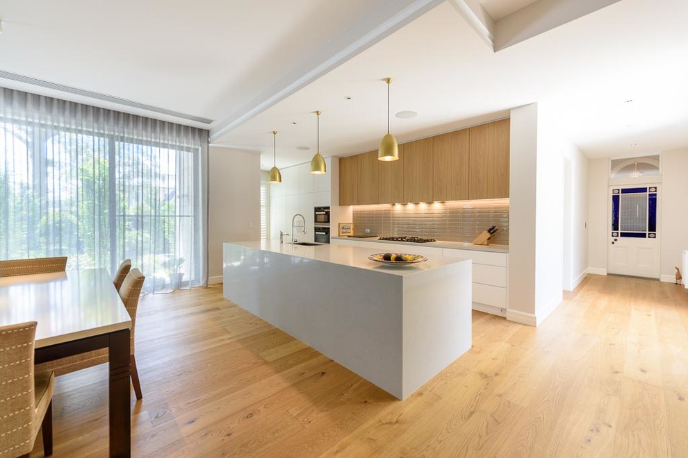 Example of a large minimalist galley light wood floor open concept kitchen design in Melbourne with an undermount sink, flat-panel cabinets, white cabinets, quartz countertops, beige backsplash, ceramic backsplash, black appliances, an island and gray countertops