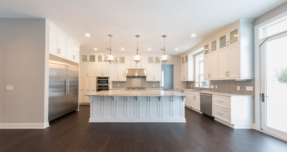 This is an example of an expansive traditional u-shaped kitchen/diner in Minneapolis with a triple-bowl sink, recessed-panel cabinets, white cabinets, granite worktops, grey splashback, glass tiled splashback, stainless steel appliances, dark hardwood flooring, an island and brown floors.