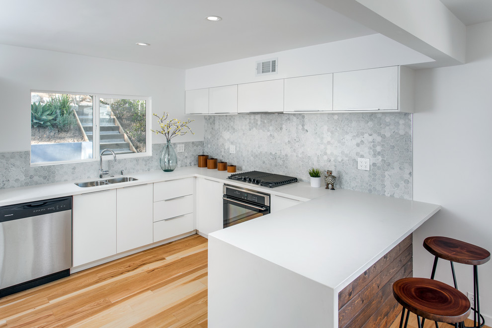 Photo of a medium sized contemporary u-shaped kitchen/diner in Los Angeles with a submerged sink, flat-panel cabinets, white cabinets, grey splashback, stainless steel appliances, light hardwood flooring, quartz worktops, mosaic tiled splashback and a breakfast bar.