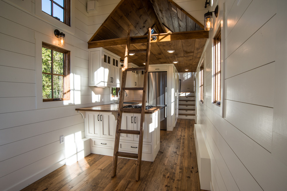 Cette photo montre une cuisine américaine nature avec un évier posé, un placard à porte shaker, des portes de placard blanches, un plan de travail en bois, un électroménager en acier inoxydable, un sol en bois brun et un sol marron.