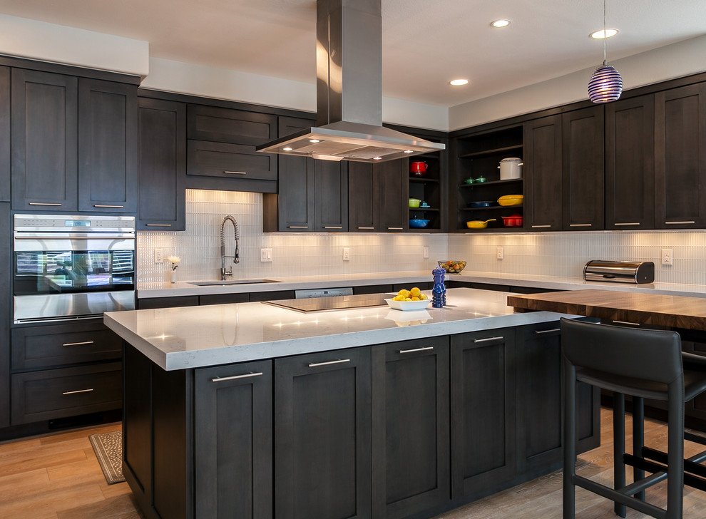 Design ideas for a large contemporary l-shaped open plan kitchen in Denver with shaker cabinets, black cabinets, white splashback, glass tiled splashback, stainless steel appliances, light hardwood flooring and an island.