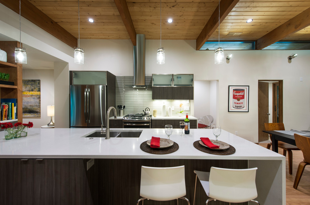 This is an example of a retro galley open plan kitchen in Denver with a submerged sink, flat-panel cabinets, dark wood cabinets, engineered stone countertops, white splashback, glass tiled splashback, stainless steel appliances, light hardwood flooring and an island.