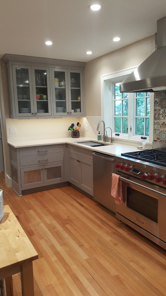 This is an example of a medium sized classic u-shaped kitchen in Other with a double-bowl sink, shaker cabinets, grey cabinets, composite countertops, white splashback, metro tiled splashback, stainless steel appliances and light hardwood flooring.