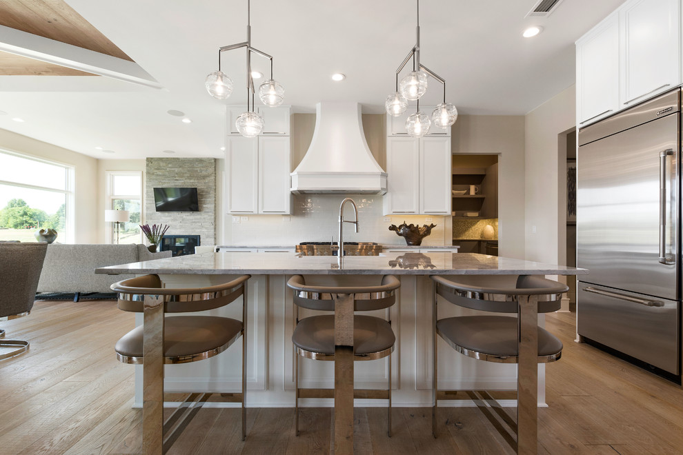 Inspiration for a transitional l-shaped light wood floor and beige floor open concept kitchen remodel in Kansas City with a farmhouse sink, recessed-panel cabinets, white cabinets, granite countertops, white backsplash, ceramic backsplash, stainless steel appliances, an island and gray countertops