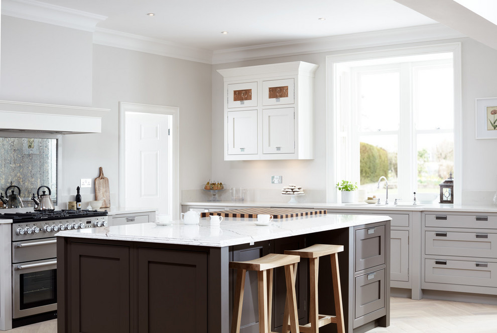 This is an example of a large traditional l-shaped kitchen in London with shaker cabinets, grey cabinets, light hardwood flooring, an island, metallic splashback, mirror splashback, stainless steel appliances, beige floors and white worktops.