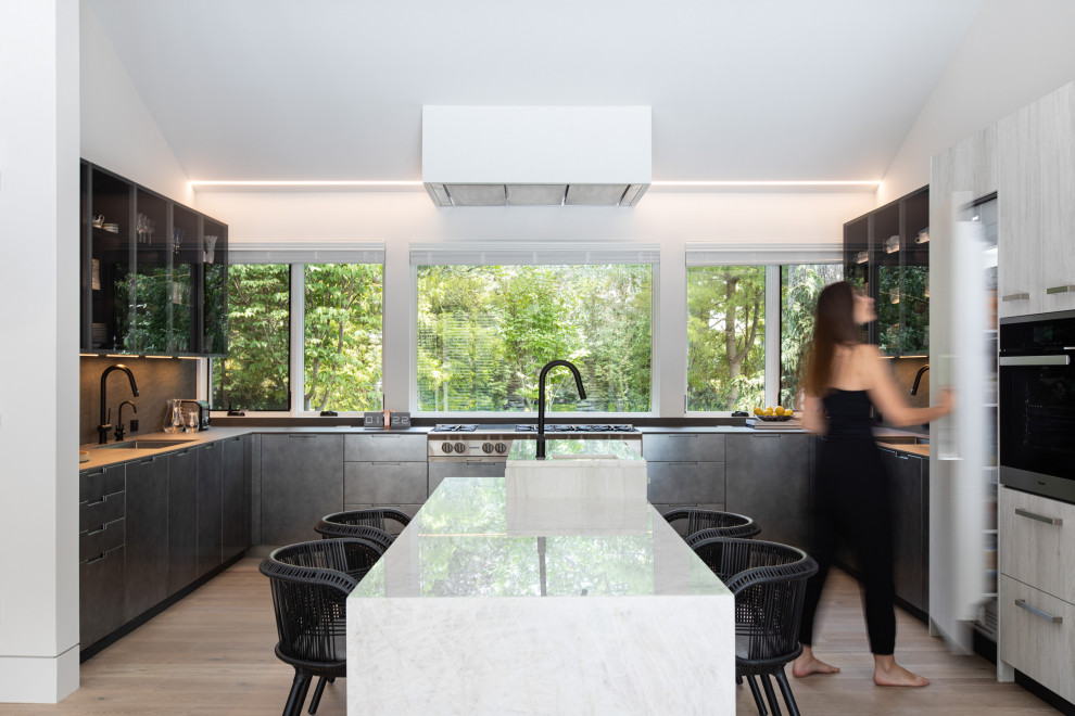Photo of a contemporary u-shaped kitchen in New York with glass-front cabinets, window splashback, light hardwood flooring, an island, beige floors and grey worktops.