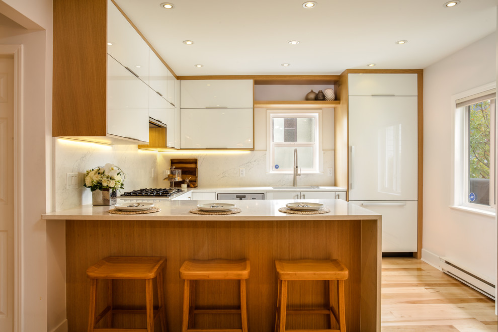 Inspiration for a medium sized contemporary u-shaped kitchen in Vancouver with a submerged sink, flat-panel cabinets, white cabinets, engineered stone countertops, white splashback, stone slab splashback, integrated appliances, light hardwood flooring, a breakfast bar and brown floors.