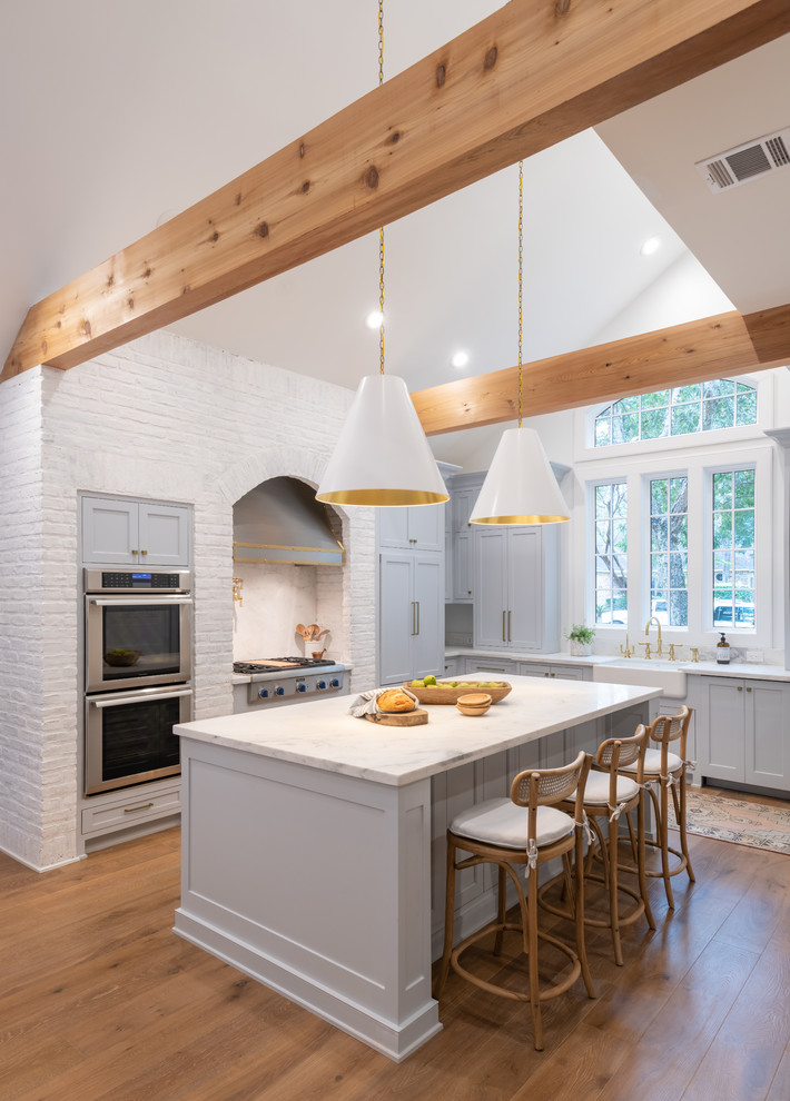 Cottage l-shaped medium tone wood floor and brown floor eat-in kitchen photo in Houston with a farmhouse sink, recessed-panel cabinets, gray cabinets, white backsplash, window backsplash, stainless steel appliances, an island and white countertops
