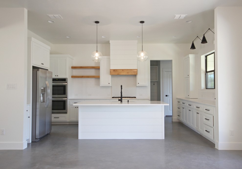 オースティンにある中くらいなカントリー風のおしゃれなアイランドキッチン (エプロンフロントシンク、シェーカースタイル扉のキャビネット、白いキャビネット、珪岩カウンター、白いキッチンパネル、サブウェイタイルのキッチンパネル、シルバーの調理設備、コンクリートの床、グレーの床、白いキッチンカウンター) の写真