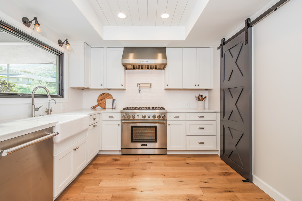 Example of a country l-shaped medium tone wood floor and brown floor kitchen design in San Diego with a farmhouse sink, shaker cabinets, white cabinets, white backsplash, stainless steel appliances, no island and white countertops