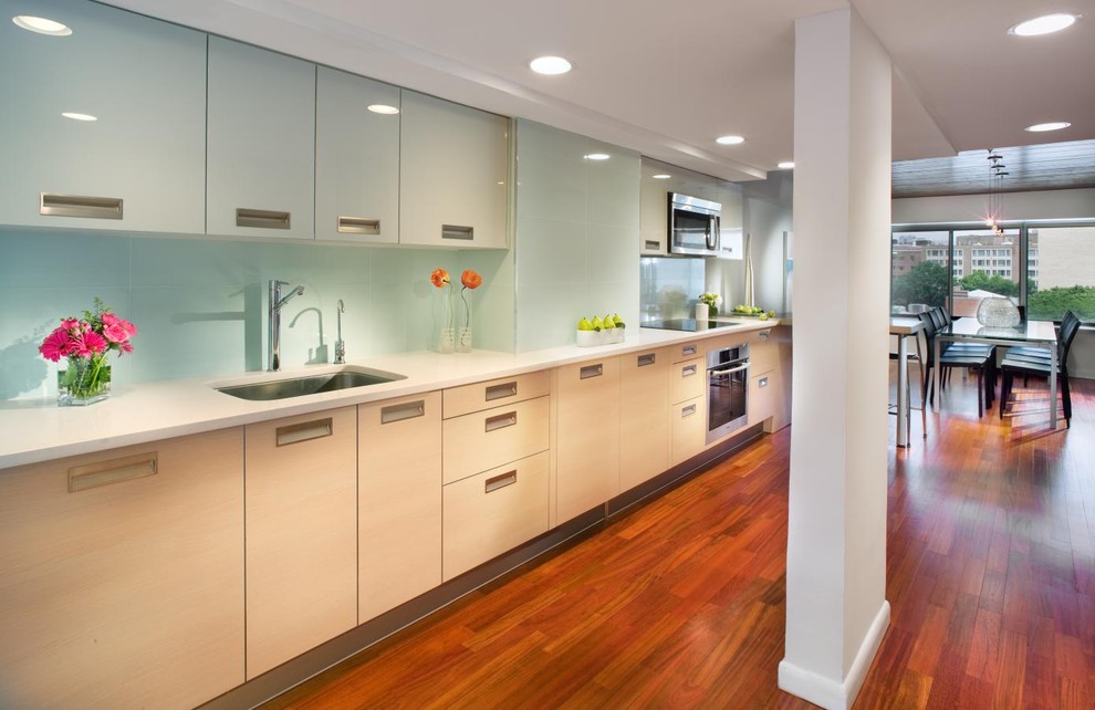Cette image montre une cuisine américaine linéaire minimaliste avec un évier encastré, un placard à porte plane, des portes de placard beiges, une crédence bleue, une crédence en feuille de verre et un électroménager en acier inoxydable.