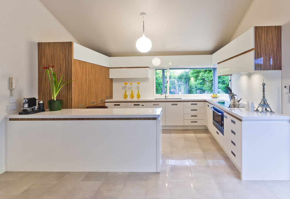 Photo of a contemporary u-shaped open plan kitchen in Los Angeles with flat-panel cabinets, white cabinets, a submerged sink, composite countertops, white splashback, glass sheet splashback, stainless steel appliances, a breakfast bar and white floors.