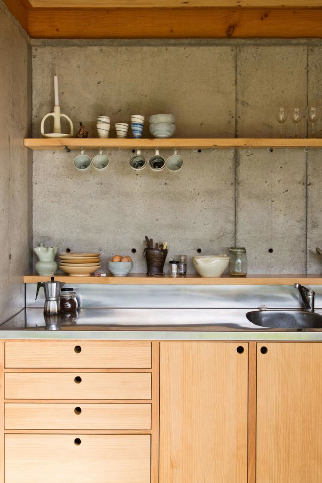 Inredning av ett kök, med en enkel diskho, luckor med infälld panel, skåp i ljust trä, bänkskiva i rostfritt stål, grått stänkskydd, rostfria vitvaror, betonggolv och en köksö