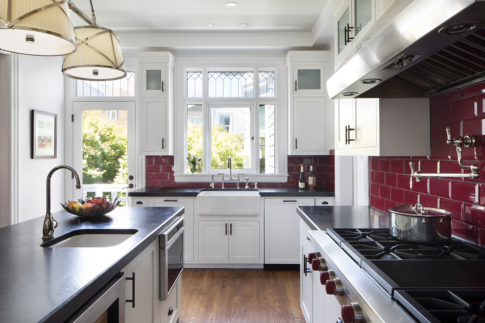 This is an example of a large victorian u-shaped enclosed kitchen in San Francisco with a belfast sink, shaker cabinets, white cabinets, red splashback, metro tiled splashback, stainless steel appliances, dark hardwood flooring, an island, brown floors and engineered stone countertops.