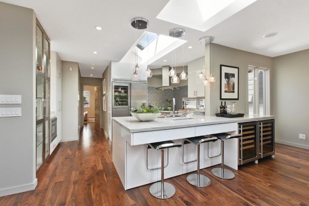 Modern inredning av ett mellanstort kök, med en undermonterad diskho, släta luckor, vita skåp, grått stänkskydd, rostfria vitvaror, mörkt trägolv, en halv köksö och brunt golv