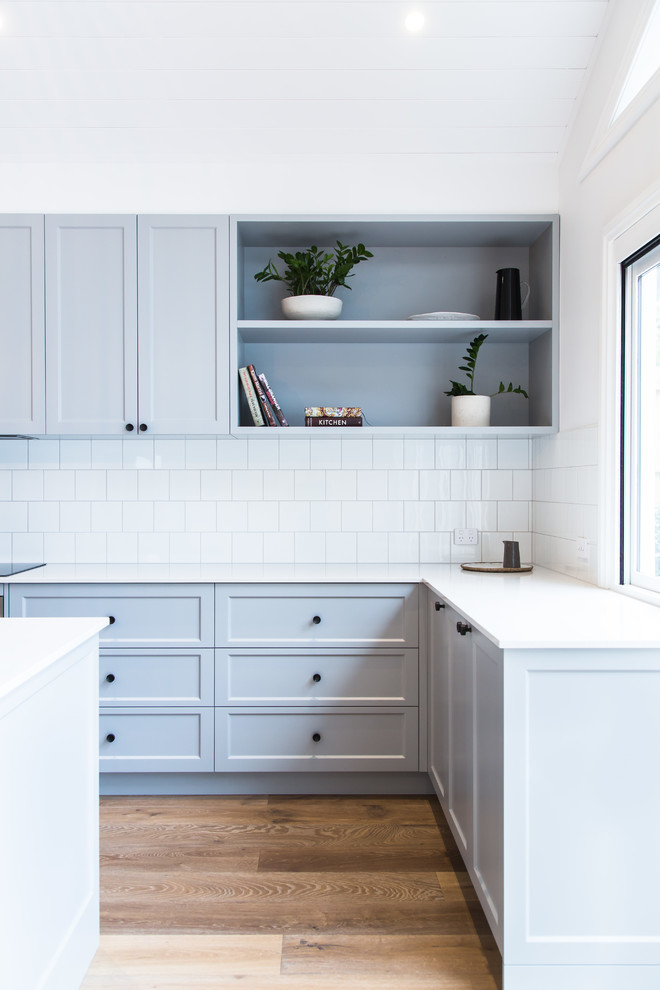 Inredning av ett modernt stort vit vitt kök, med en undermonterad diskho, skåp i shakerstil, grå skåp, bänkskiva i kvarts, vitt stänkskydd, stänkskydd i keramik, svarta vitvaror, mellanmörkt trägolv, en köksö och brunt golv
