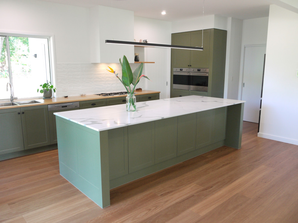 This is an example of a medium sized contemporary galley kitchen/diner in Sunshine Coast with a double-bowl sink, recessed-panel cabinets, green cabinets, engineered stone countertops, white splashback, ceramic splashback, stainless steel appliances, vinyl flooring, an island, brown floors and white worktops.