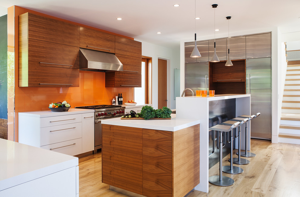 Bild på ett funkis u-kök, med släta luckor, skåp i mellenmörkt trä, orange stänkskydd, glaspanel som stänkskydd, rostfria vitvaror, ljust trägolv och en köksö