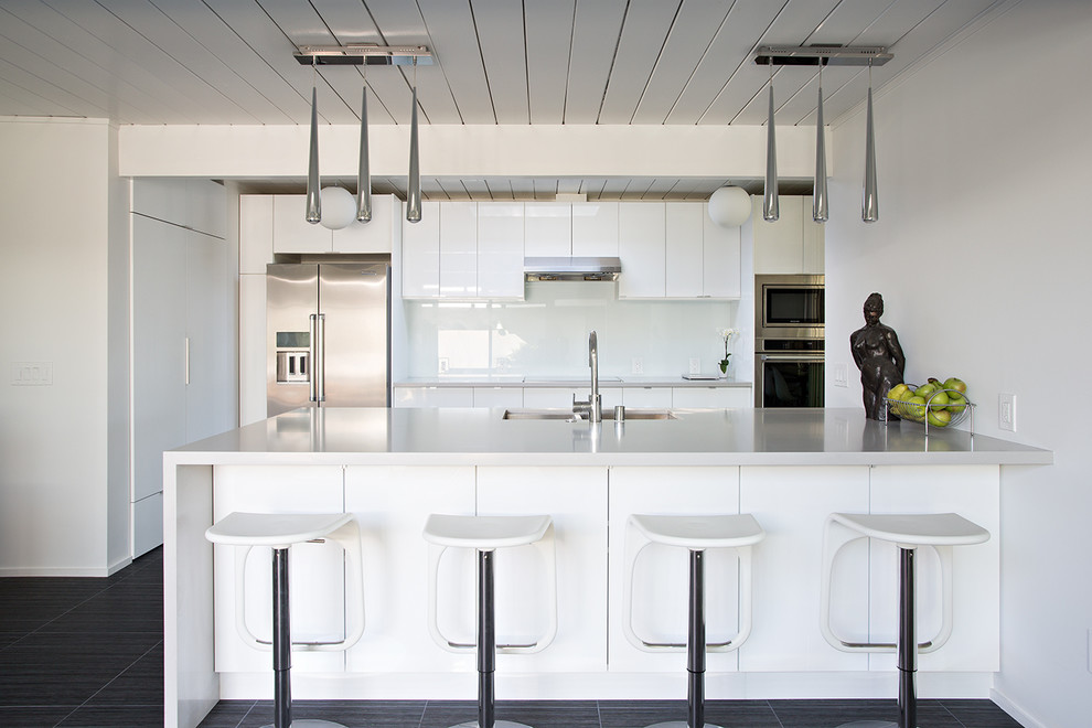 Exemple d'une cuisine rétro avec un placard à porte plane, des portes de placard blanches, une crédence blanche, une crédence en feuille de verre, un électroménager en acier inoxydable et une péninsule.