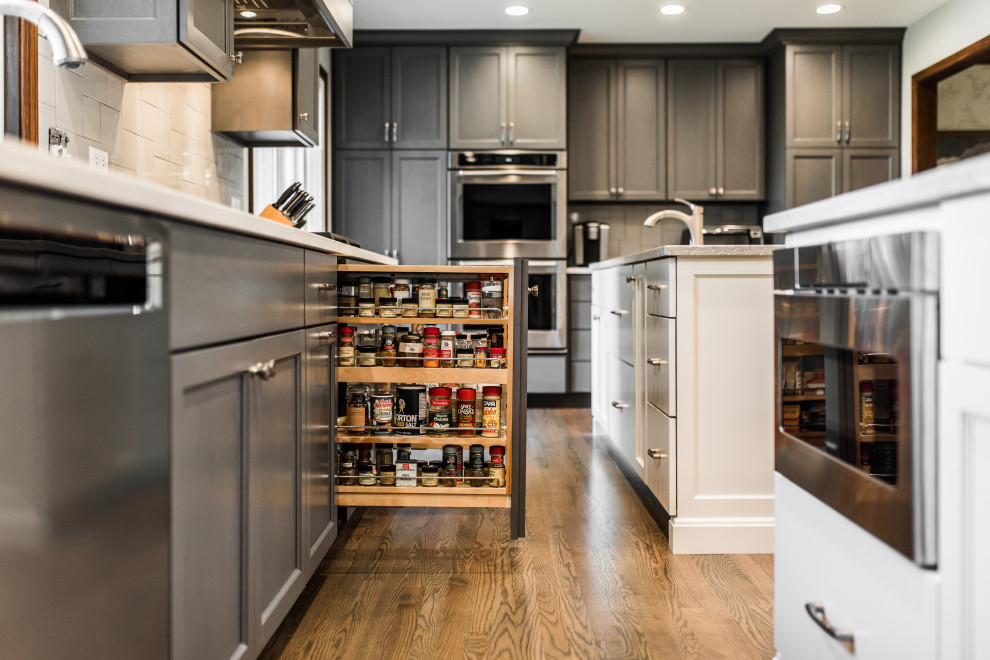 This is an example of a large classic galley kitchen/diner in Chicago with a double-bowl sink, shaker cabinets, grey cabinets, engineered stone countertops, white splashback, metro tiled splashback, stainless steel appliances, medium hardwood flooring, multiple islands, brown floors and white worktops.