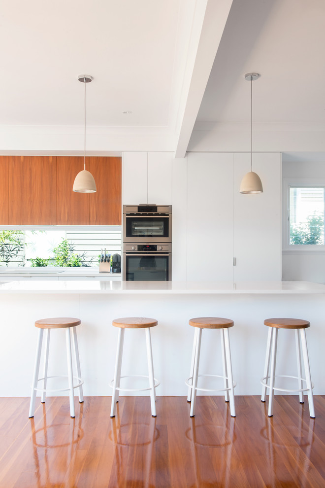 Zweizeilige, Mittelgroße Moderne Küche mit flächenbündigen Schrankfronten, weißen Schränken, Kücheninsel, Küchengeräten aus Edelstahl und braunem Holzboden in Brisbane