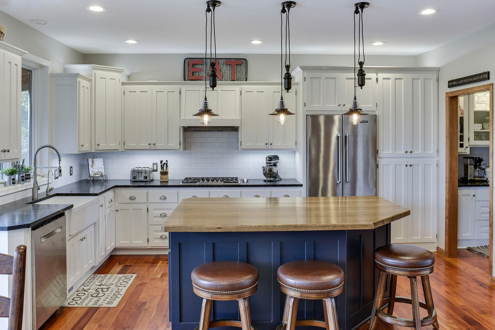 This is an example of a classic l-shaped kitchen/diner in Minneapolis with a belfast sink, shaker cabinets, blue cabinets, white splashback, metro tiled splashback, stainless steel appliances, medium hardwood flooring and an island.