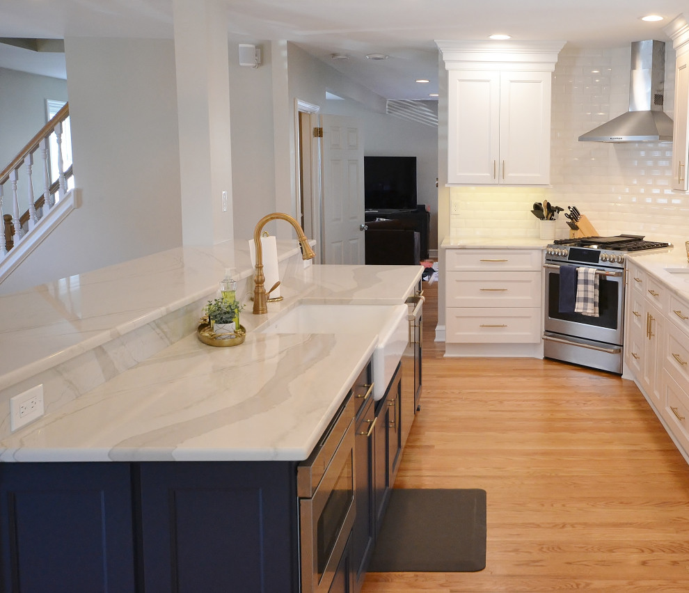 Example of a huge transitional l-shaped light wood floor, brown floor and exposed beam eat-in kitchen design in Philadelphia with a farmhouse sink, recessed-panel cabinets, blue cabinets, marble countertops, white backsplash, subway tile backsplash, stainless steel appliances, a peninsula and white countertops