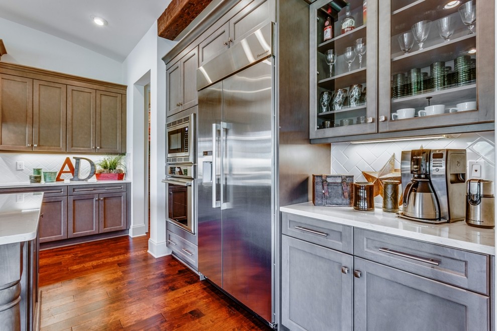 This is an example of a large mediterranean l-shaped open plan kitchen in Jacksonville with a belfast sink, shaker cabinets, grey cabinets, engineered stone countertops, white splashback, ceramic splashback, stainless steel appliances, medium hardwood flooring, an island and brown floors.