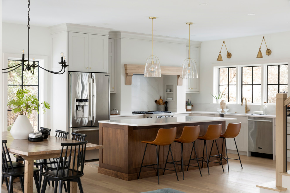 Example of a transitional l-shaped light wood floor and beige floor kitchen design in Minneapolis with an undermount sink, shaker cabinets, white cabinets, white backsplash, stone slab backsplash, stainless steel appliances, an island and white countertops