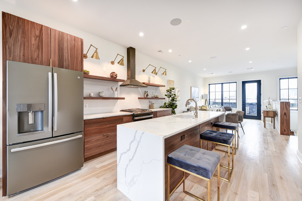 Example of a mid-sized galley light wood floor and brown floor open concept kitchen design in DC Metro with a single-bowl sink, flat-panel cabinets, medium tone wood cabinets, quartz countertops, white backsplash, ceramic backsplash, stainless steel appliances, an island and white countertops