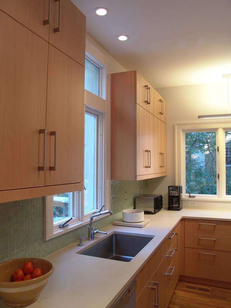 Modern inredning av ett kök, med en undermonterad diskho, släta luckor, skåp i ljust trä, grönt stänkskydd, stänkskydd i mosaik och rostfria vitvaror