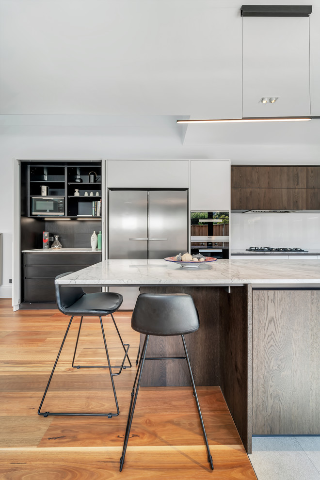 Photo of a large modern l-shaped kitchen/diner in Sydney with a double-bowl sink, flat-panel cabinets, white cabinets, marble worktops, white splashback, glass sheet splashback, stainless steel appliances, medium hardwood flooring, an island, brown floors and grey worktops.