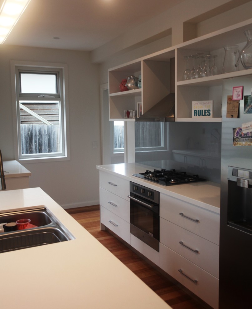 Photo of a modern galley kitchen/diner in Melbourne with a submerged sink, white cabinets, grey splashback, glass sheet splashback, stainless steel appliances, medium hardwood flooring and an island.