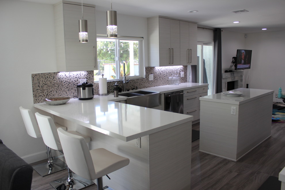 Example of a mid-sized transitional u-shaped porcelain tile and brown floor eat-in kitchen design in Los Angeles with a farmhouse sink, flat-panel cabinets, white cabinets, solid surface countertops, multicolored backsplash, mosaic tile backsplash, stainless steel appliances and an island